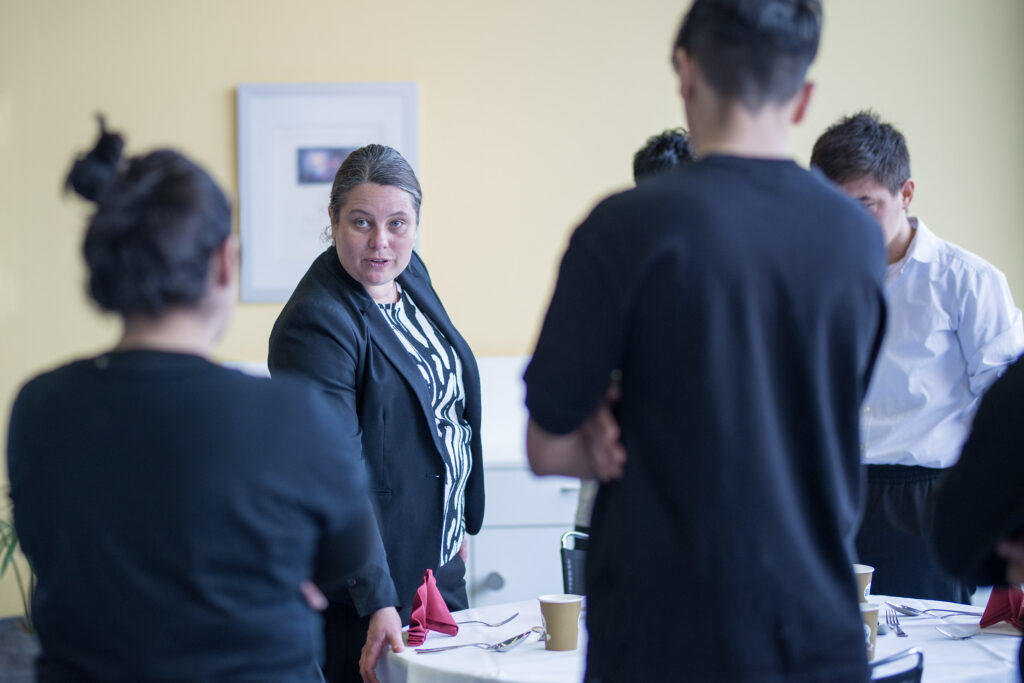 Die Lehrperson erklärt den Lernenden: Genauigkeit im Service, auch das Besteck hat seinen festen Platz: Eine Daumenbreite von der Kante entfernt.