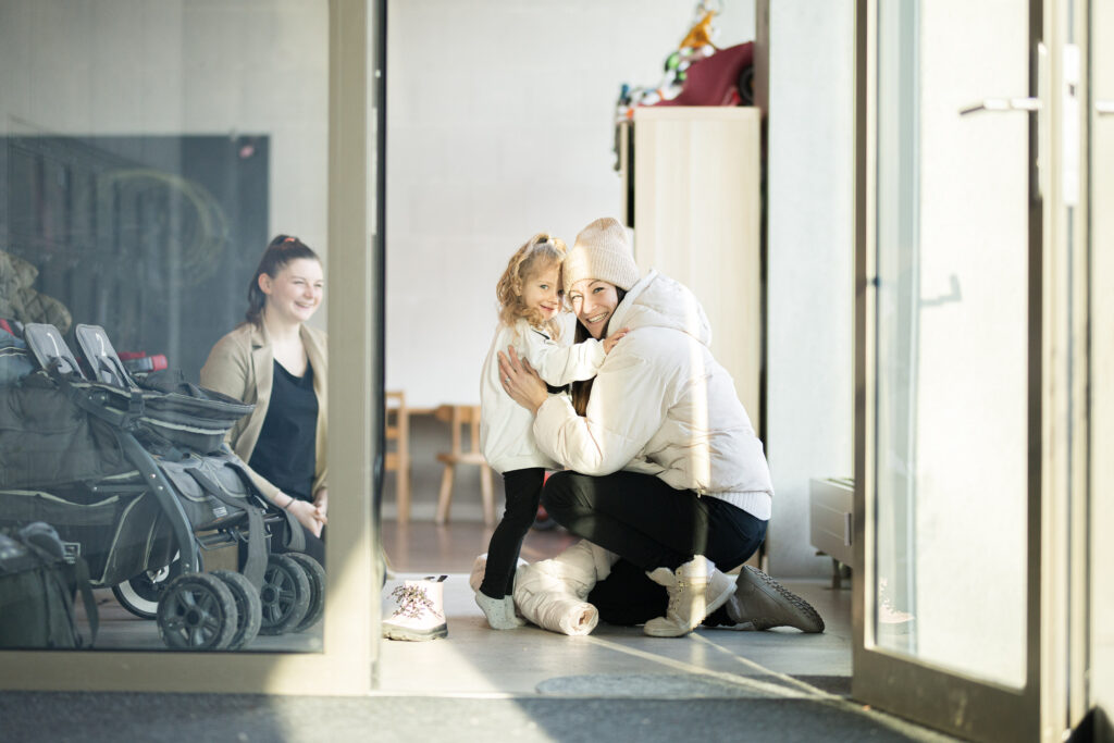 Eine Frau verabschiedet sich morgens vor einer Kindertagesstätte von ihrem kleinen Kind.