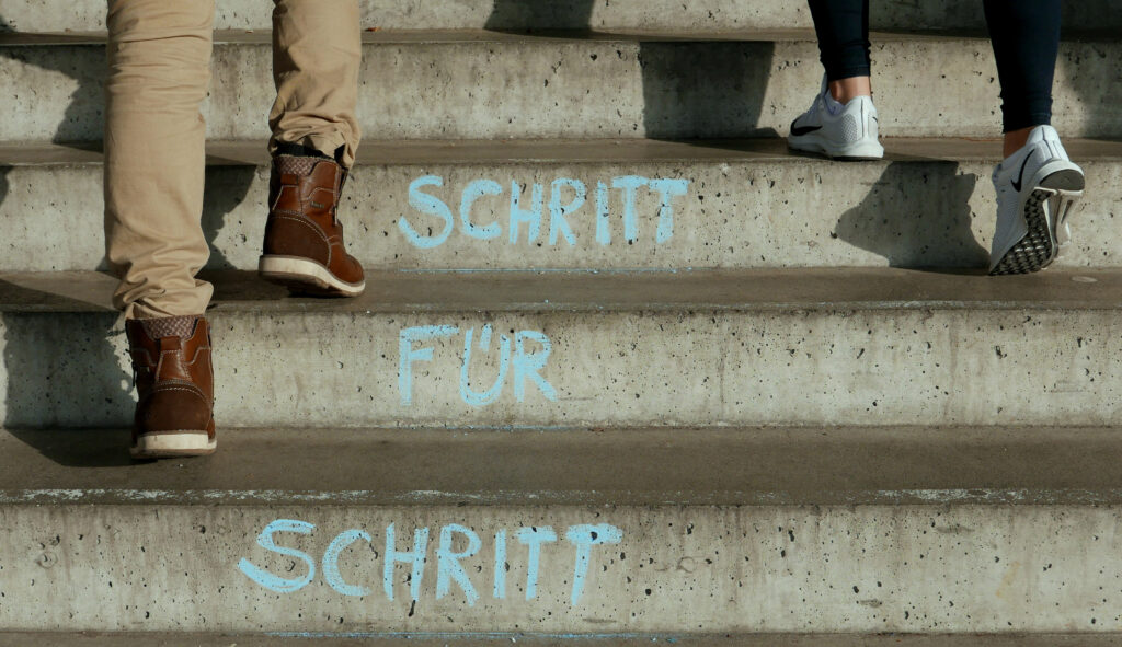 Das Bild zeigt eine Treppe aus Beton. Es sind die Füsse von zwei Personen ersichtlich, die die Treppe hochsteigen. Auf den Stirnseiten der Treppe steht in blauer Schrift «Schritt für Schritt»